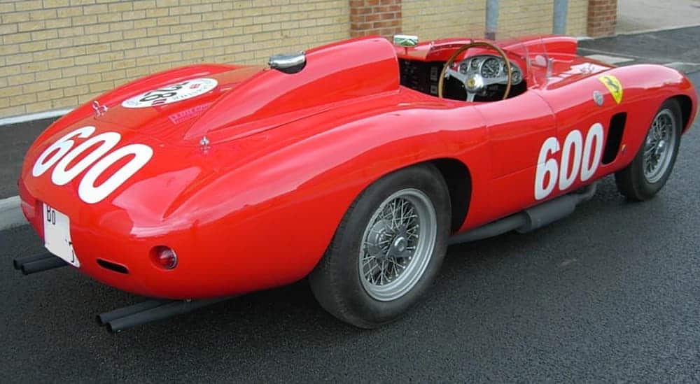 A rear look of a red 1956 Ferrari 290 MM that's parked next to a brick building is shown. 