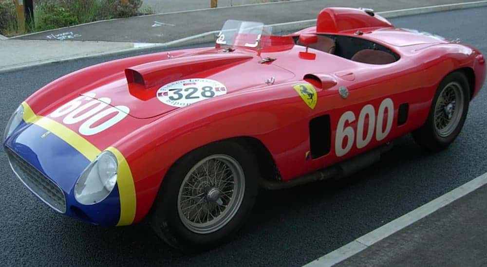 A red, with race number decals on it, 1956 Ferrari 290 MM is parked in an empty parking lot. 