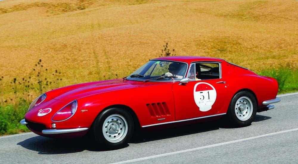 A red 1964 Ferrari 275 GTB/C Speciale is driving past a hay field on a sunny day. 