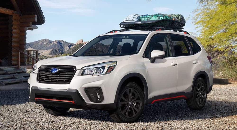 A white 2019 Subaru Forester is parked in front of a log cabin with mountains in the distance. 