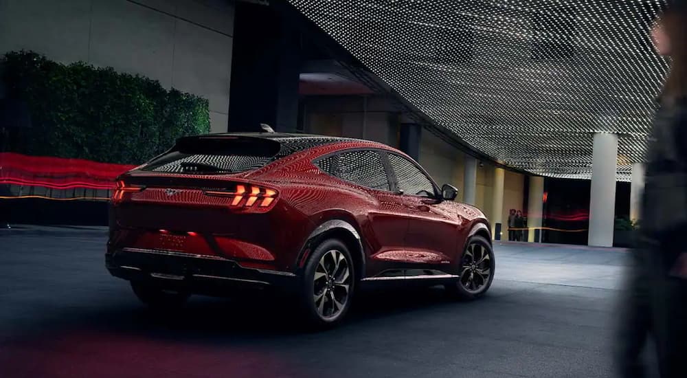 A red 2021 Mach-E, which wins when comparing the 2021 Ford Mustang Mach-E vs Tesla Model Y, is parked under accent lights in front of a city building at night. 