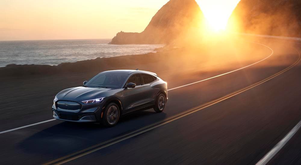 A grey 2021 Ford Mustang Mach-E, which is a popular subject in current auto news, is driving on a road next to an ocean during a sunset. 
