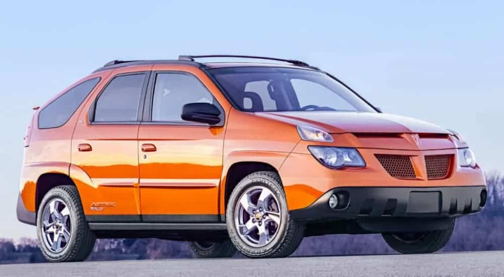 An orange 2004 Pontiac Aztek is parked in an empty parking lot with mountains in the distance. 