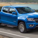 A blue 2020 Chevy Colorado, which wins when comparing the 2020 Chevy Colorado vs 2020 Ford Ranger, is driving on a grass lined road next to a lake.