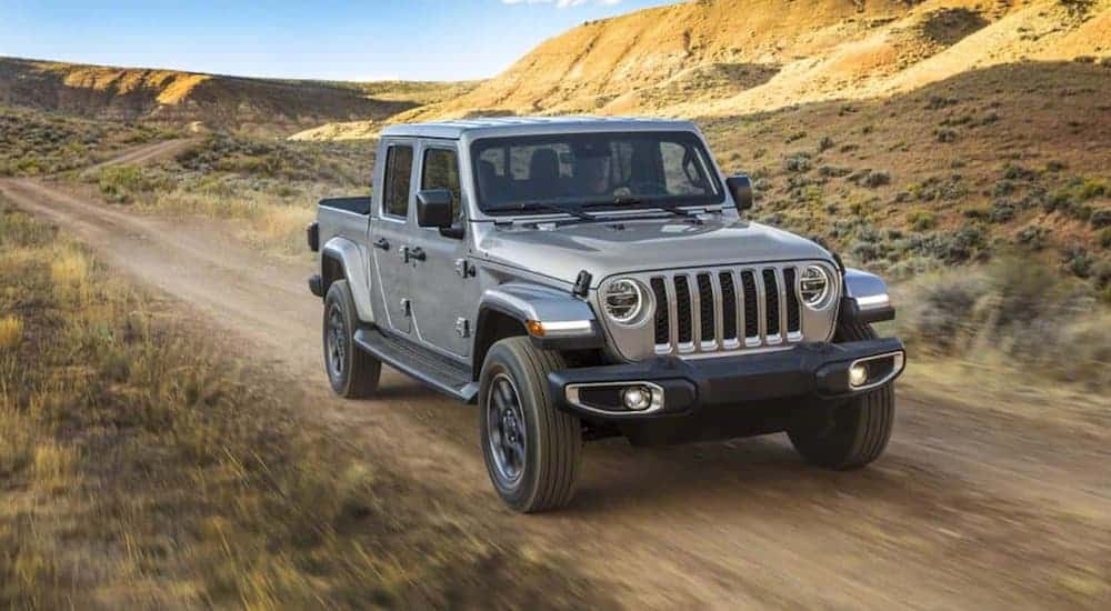 A silver 2020 Jeep Gladiator is driving on a dirt trail with mountains in the distance. 