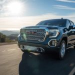 A dark grey 2020 GMC Sierra 1500 Denali is driving on a sunny highway with mountains in the distance.