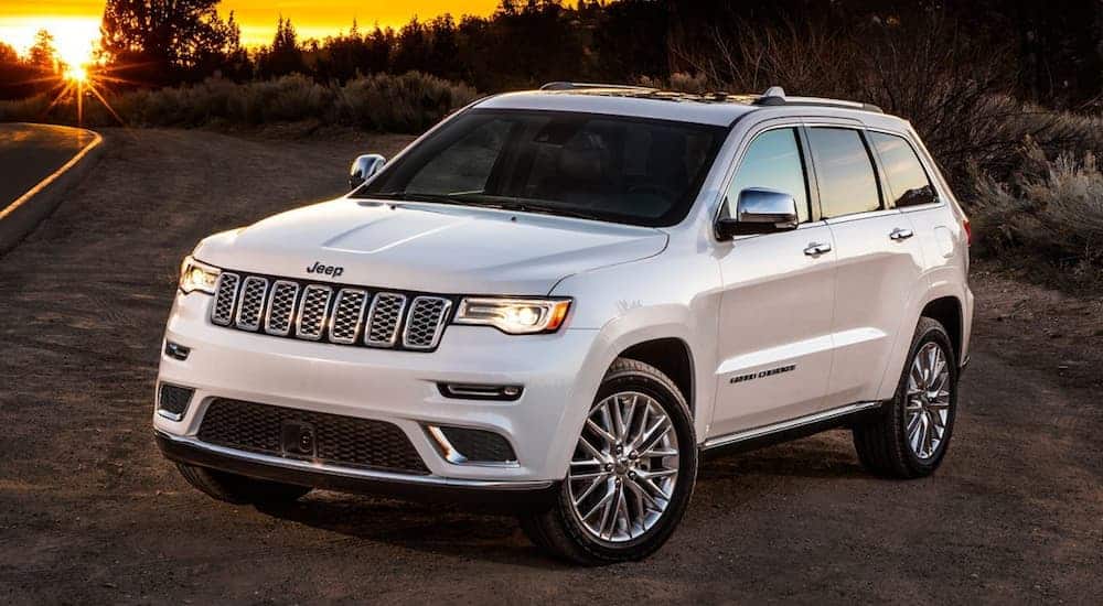 A white 2018 Jeep Grand Cherokee, which is popular among used Jeeps for sale, is parked on the side of a road next to bushes with the sun setting in the distance.