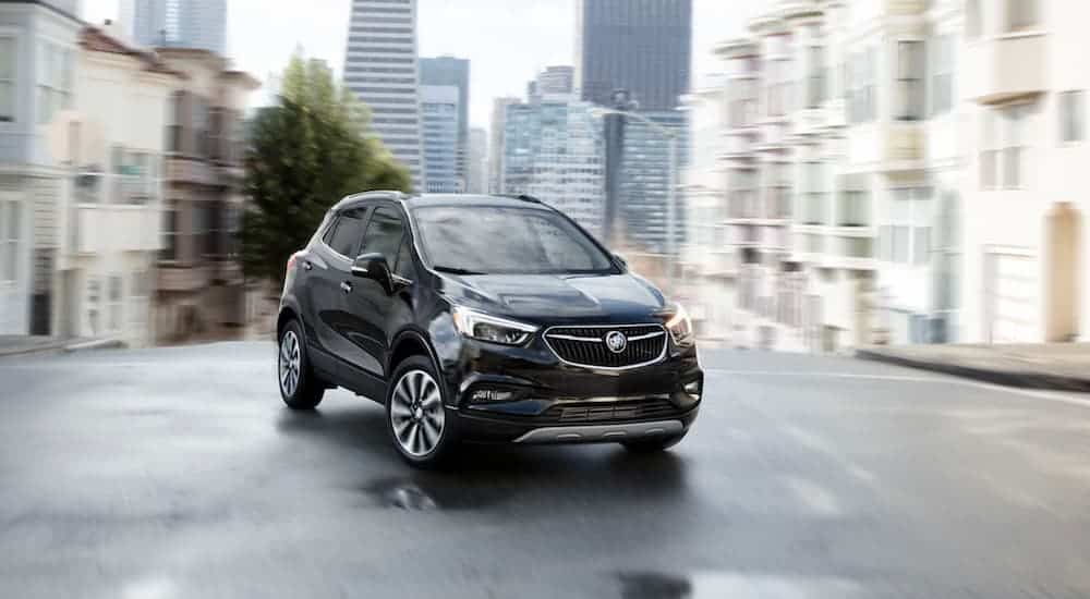 A black 2020 Buick Encore is driving on a city street with city buildings in the distance. 