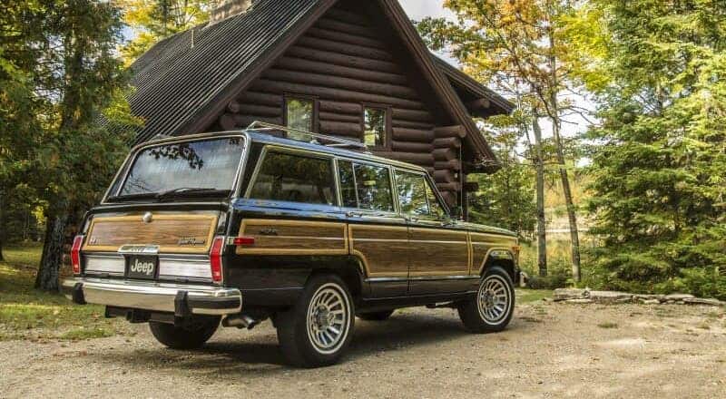 A black and wood-paneled Jeep Grand Wagoner is parked in front of a log cabin.