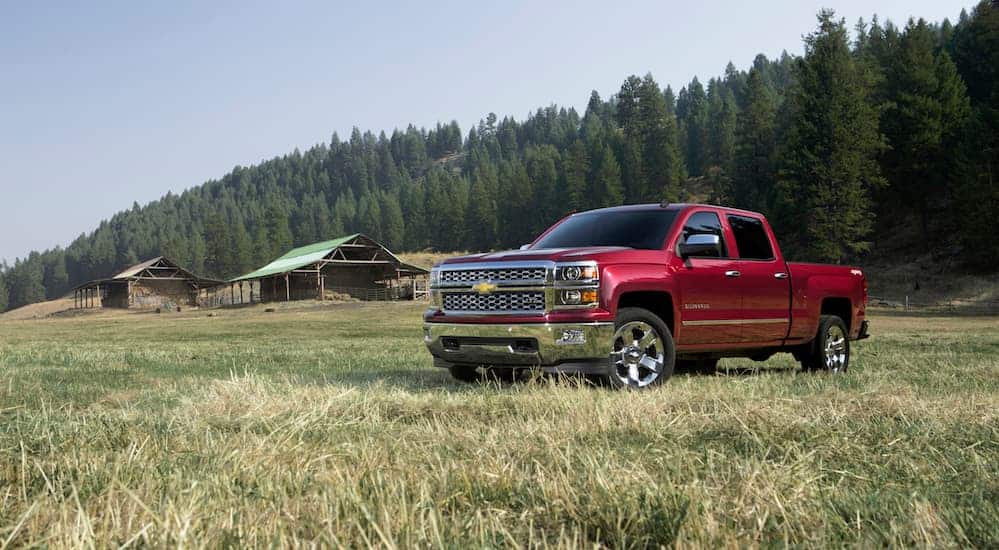 A red 2014 Chevy Silverado 1500 is parked in a grassy field with a log cabin in the distance. 