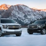 A 2015 Chevy Suburban is parked next to a 2015 Tahoe in the snow in front of mountains.