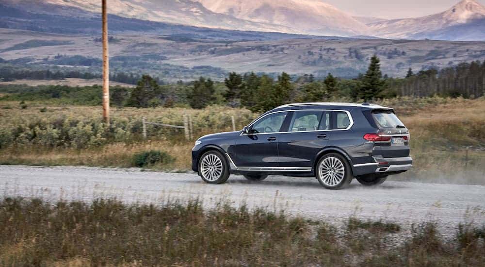 A blue 2020 BMW X7 is shown from the side driving on a dirt road in front of distant mountains.