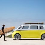 A surfer is walking towards a yellow and white 2022 VW Microbus prototype that is parked on a beach.