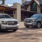 A grey 2020 Chevy Suburban and a white 2020 Chevy Tahoe are parked in front of a large home.