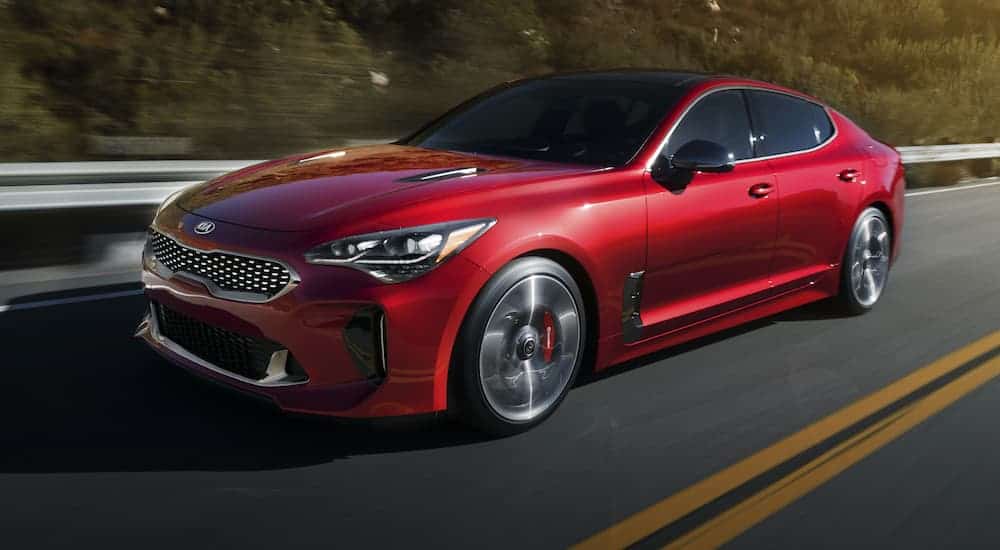 A red 2020 Kia Stinger is driving past a guardrail and blurred trees.