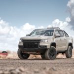 A tan 2021 Chevy Colorado ZR2 is parked in the desert as a red special edition races behind it.