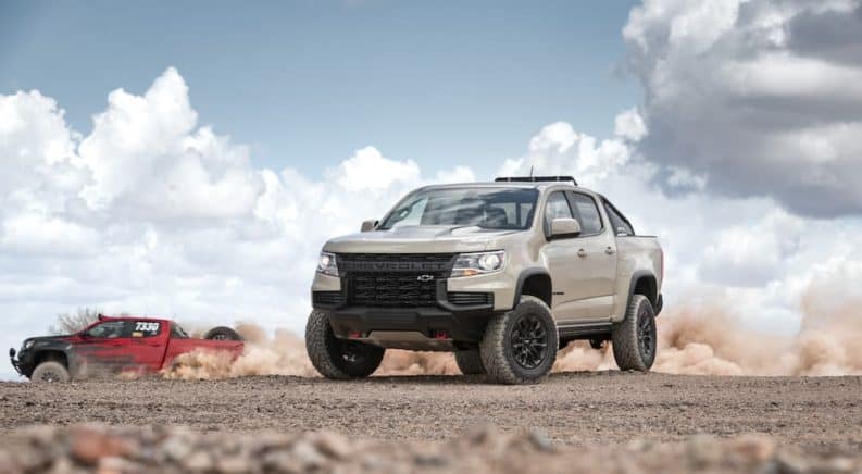 A tan 2021 Chevy Colorado ZR2 is parked in the desert as a red special edition races behind it.