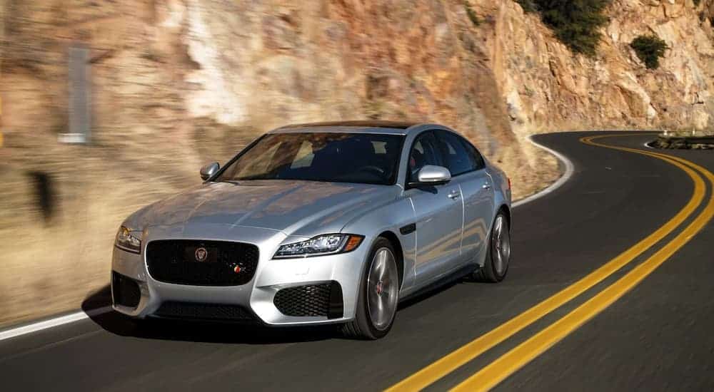 A silver 2016 Jaguar XF is driving past rock face on a winding road after leaving a used car dealer.