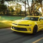 A yellow 2017 Chevy Camaro, which is popular among used cars for sale, is driving on a sunny road past trees.