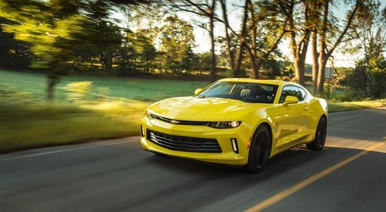 A yellow 2017 Chevy Camaro, which is popular among used cars for sale, is driving on a sunny road past trees.