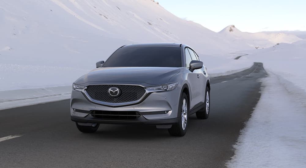 A silver 2020 Mazda CX-5 is driving on a road in front of snow-covered hills.