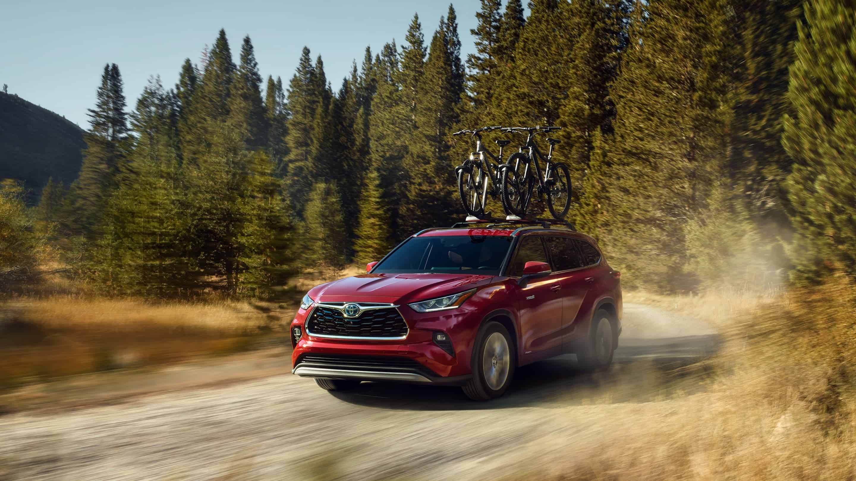 A red 2020 Toyota Highlander is driving on a dirt trail in front of trees.