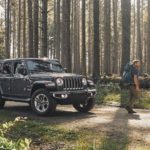 A couple is walking away from a gray 2020 Jeep Wrangler Unlimited, from a local Jeep dealership, that is parked in the woods.