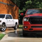 Two Ram trucks, a white and a red 2020 Ram 1500, are parked in front of a wooden garage.