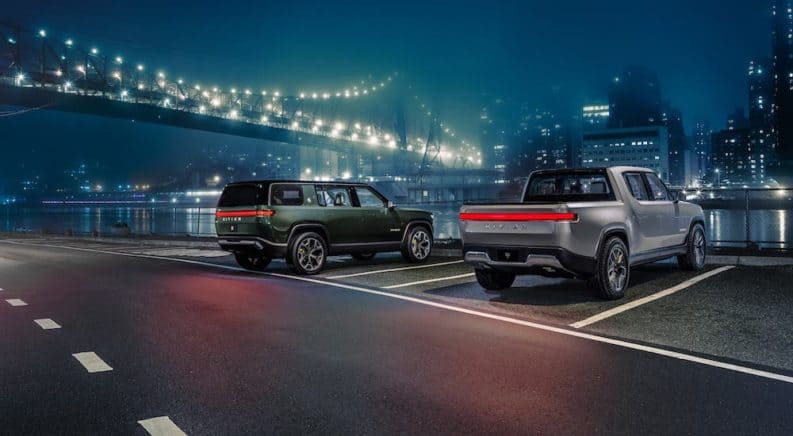 A green Rivian R1S and silver R1T are parked in front of a city bridge at night.