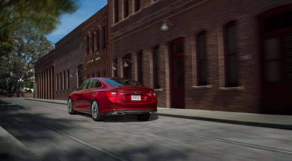 A red 2020 Chevy Malibu RS is driving past a red brick building after winning the 2020 Chevy Malibu vs 2020 Toyota Camry comparison.
