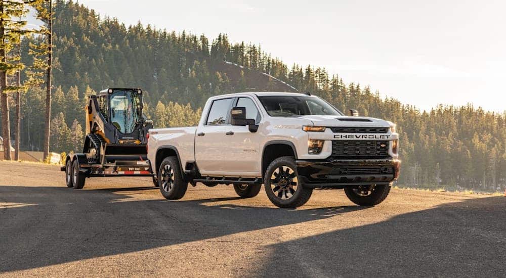 A white 2020 Chevy Silverado 2500HD is parked on a mountain highway and towing a trailer with a skid steer.