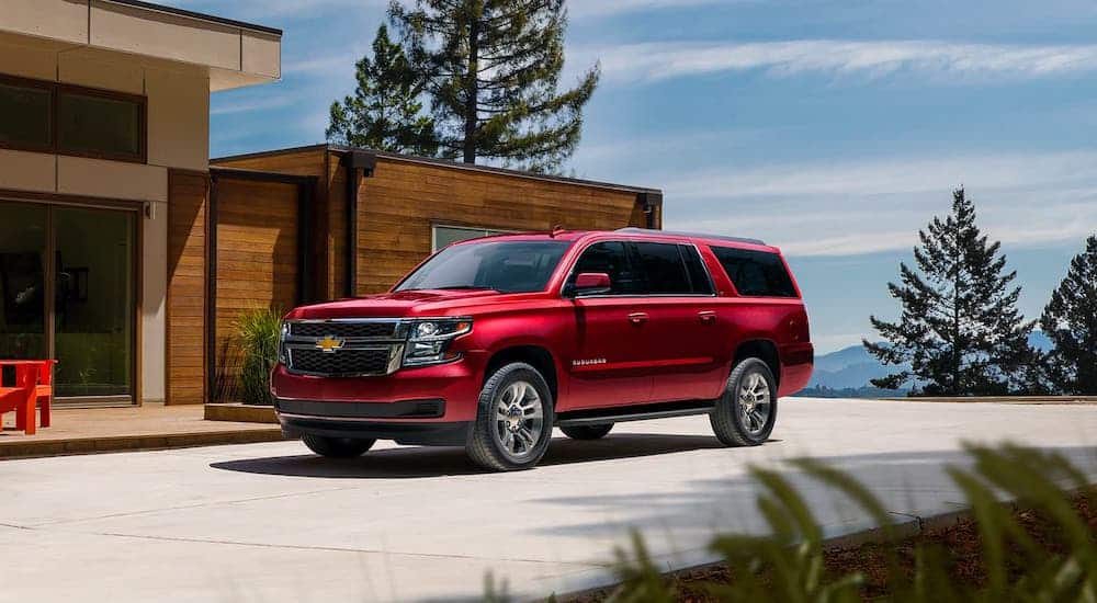 A red 2020 Chevy Suburban is parked in front of a modern cabin with mountains in the distance.