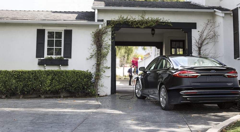 A black 2020 Ford Fusion Hybrid is being charged in front of a white house after winning the 2020 Ford Fusion vs 2020 Mazda 6 comparison.