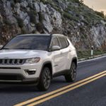 A white 2020 Jeep Compass is driving on a highway in front of a rock face.