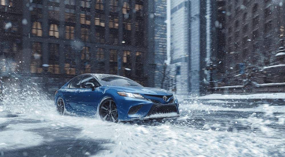 A blue 2020 Toyota Camry is driving on a snowy city road.