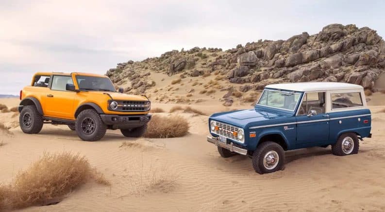 A yellow 2021 Ford Bronco 2door is parked in sand next to a blue 1970s Bronco.