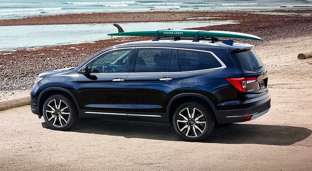 One of the newer Honda SUVs, a blue 2021 Honda Pilot, is parked at a beach with a surfboard on the roof rack.