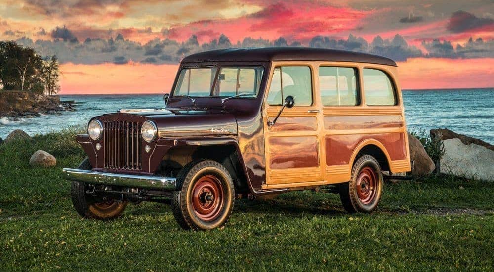 A red wood-paneled 1940s Jeep Willys Wagon is parked on grass in front of a vibrant pink sky.
