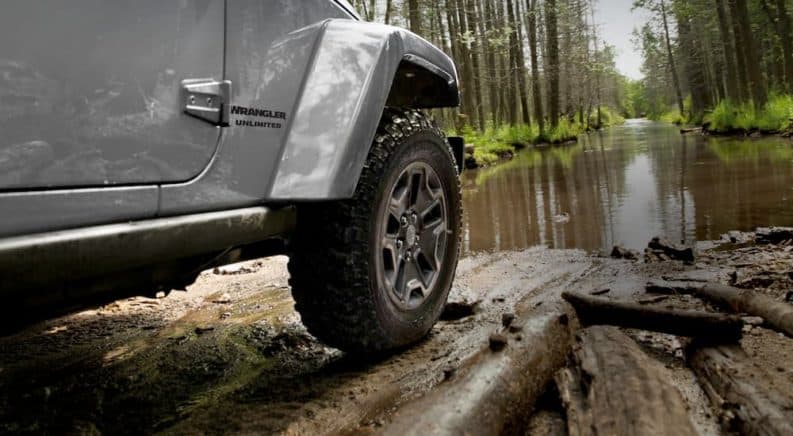 A closeup of the fender of a silver 2017 Jeep Wrangler Unlimited is shown in front of a muddy trail.