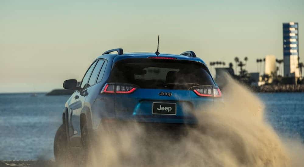 A blue 2020 Jeep Cherokee Trailhawk Elite is driving in the sand at the beach, shown from the rear.