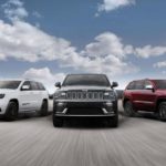 Five different trims of the 2020 Jeep Grand Cherokee are driving in a line with a blue sky and clouds above.