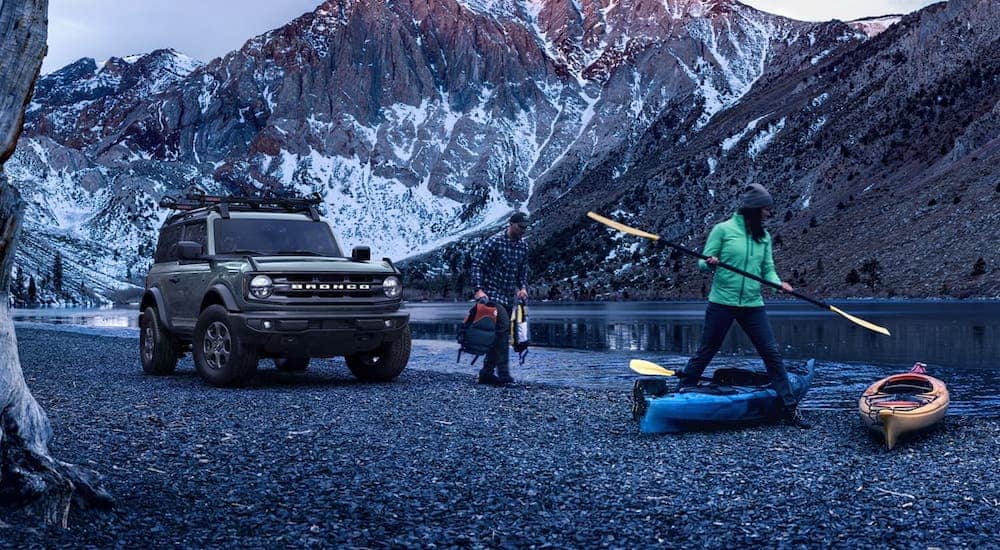 People are preparing kayaks next to a river and a gray 2021 Ford Bronco 2-door with snowy mountains in the distance.
