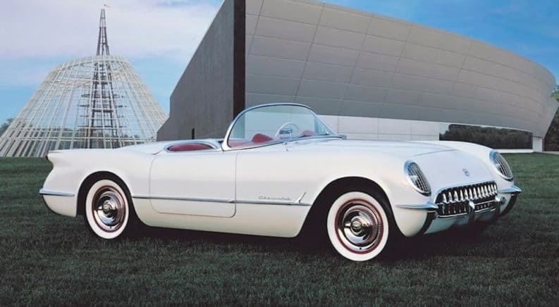 A white convertible 1953 Chevy Corvette, which would be hard to find at a Chevy dealer, is parked in front of a large grey building.