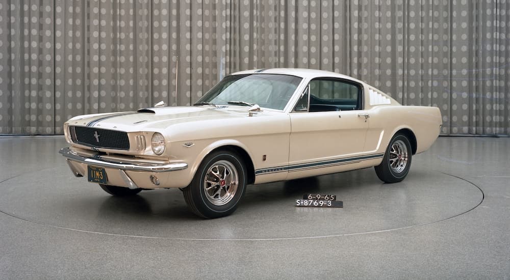 A tan 1965 Ford Mustang Fastback is displayed in a showroom.
