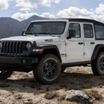 One of the classic Jeep SUVs, a white 2020 Jeep Wrangler Willys Edition, is parked in front of a lake and mountains.