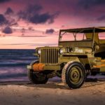 A 1940s Jeep Willys is parked on a beach in front of a vibrant purple and pink sunset.