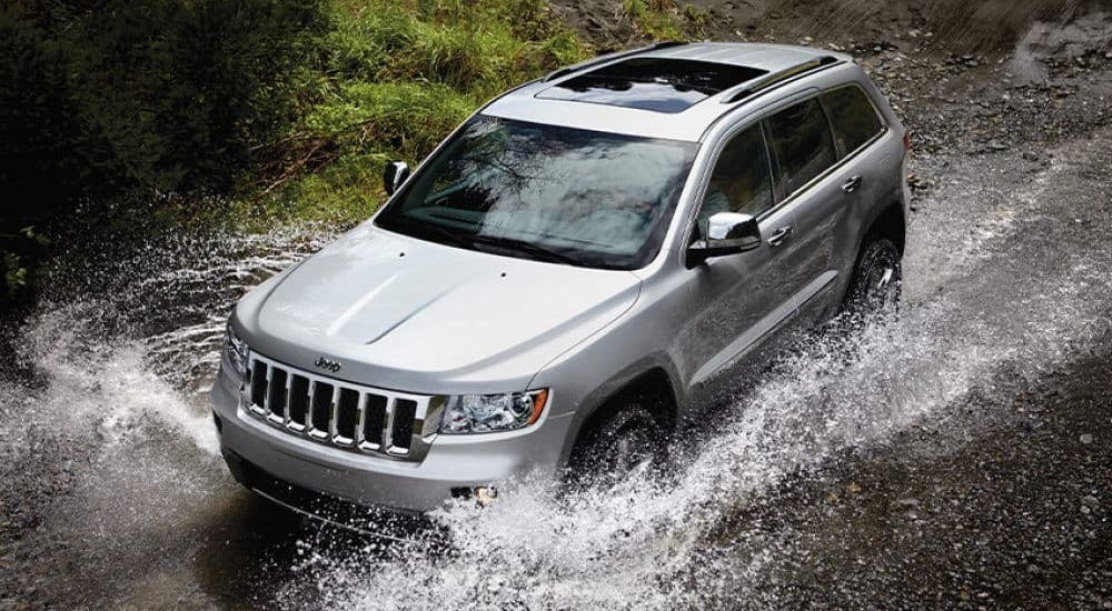 A common used car for sale near you, a silver 2014 Jeep Grand Cherokee, is driving through a puddle.