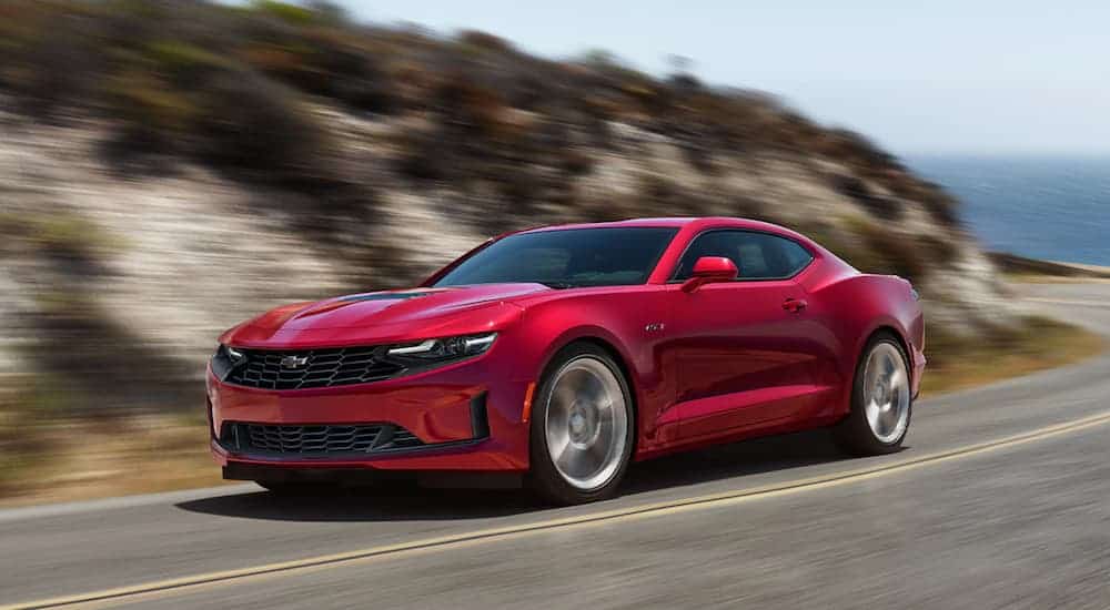 A 2021 Chevy Camaro in Wild Cherry red is driving past a small hill with shrubs.