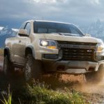 A tan 2021 Chevy Colorado ZR2 is driving on a dirt path in front of mountains.