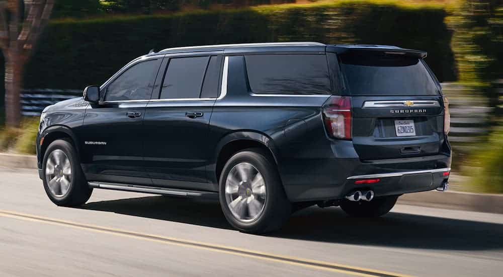 A black 2021 Chevy Suburban is driving past a fence and shrubs during a 2021 Chevy Suburban vs 2021 Cadillac Escalade comparison.